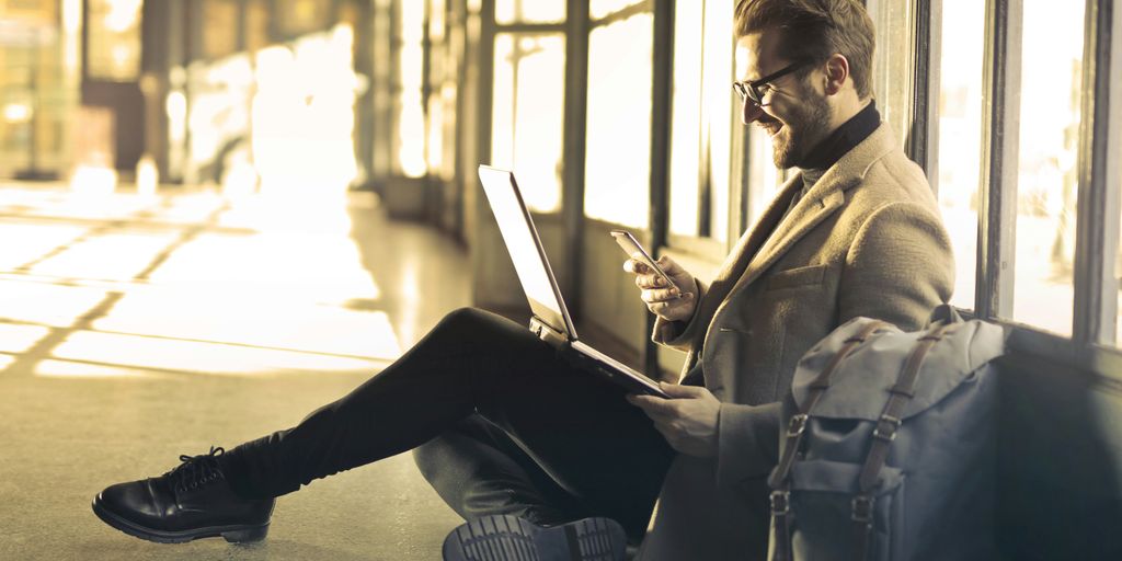 man sitting near window holding phone and laptop